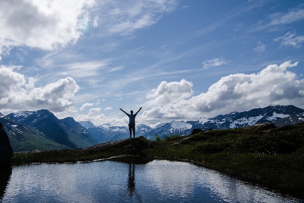 Mountain scenary with clouds in the sky, and a person stood next to a lake 