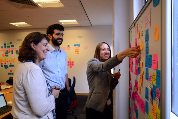 Three colleagues standing at a wall having a meeting, writing their ideas on post-it notes and sticking the notes to the wall. 