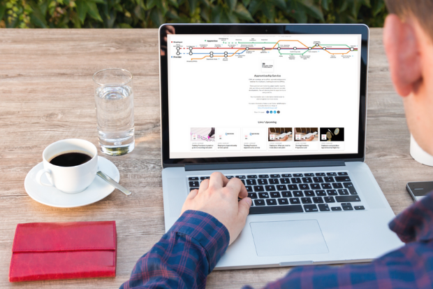 Man sitting at table with his left hand on laptop which is open and the screen displayed is that of the Apprenticeship services webinar programme. On the left there is also a cup of black coffee , glass of water and a red wallet on the table .